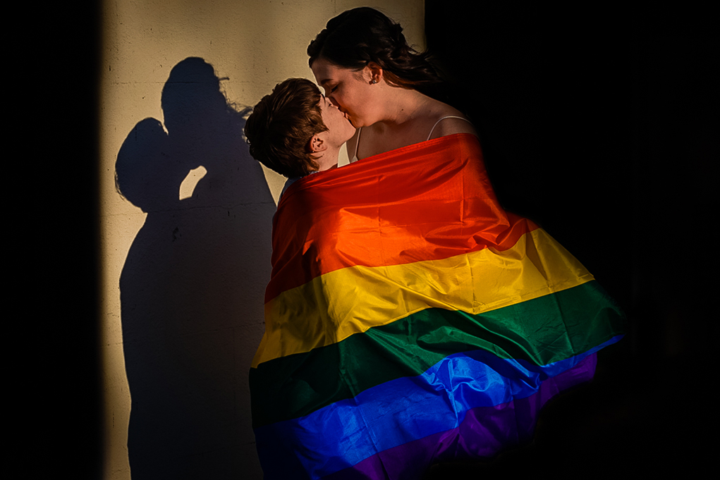 Two brides kissing with their pride flag during sunset during their Great Falls engagement photos by Potok's World Photography, engagement photographers in Maryland.